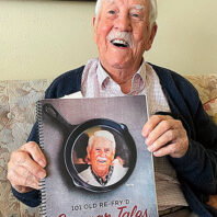An older man sits on a couch holding a spiral-bound book titled “101 Old Re-Fry’d Cougar Tales,” which features a photo of the same person on the cover inside a cast-iron skillet graphic.