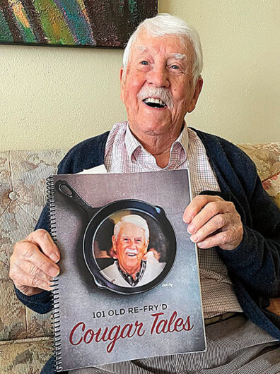 An older man sits on a couch holding a spiral-bound book titled “101 Old Re-Fry’d Cougar Tales,” which features a photo of the same person on the cover inside a cast-iron skillet graphic.