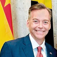 Portrait crop of a man wearing a blue suit, white shirt, and red tie, standing in front of colorful international flags.