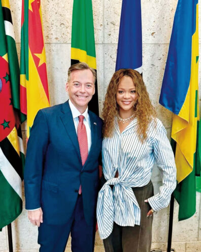 A man and woman pose for a photo in front of a row of international flags inside a government or institutional building. One person wears a business suit, and the other wears a striped blouse and necklace.