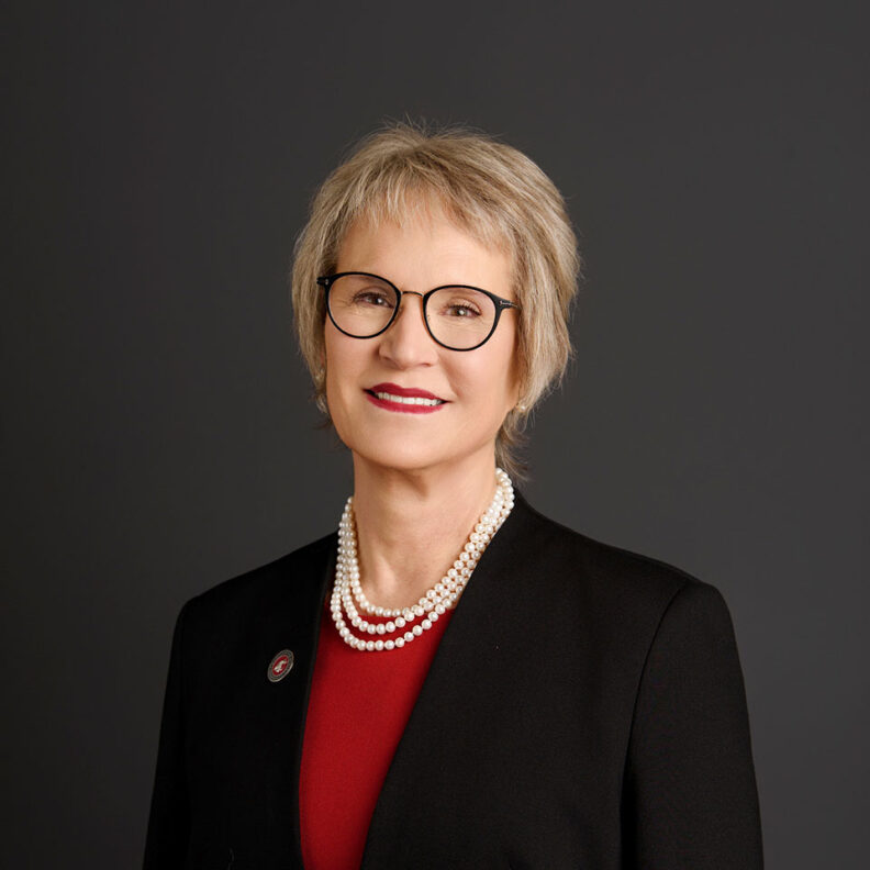 Studio portrait of a woman wearing glasses, a black blazer, a red top, and a pearl necklace against a dark gray background.
