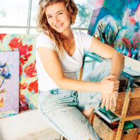 Woman seated on a stool in an art studio surrounded by colorful paintings, paintbrushes, and artist supplies.