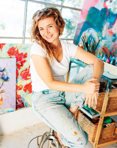 Woman seated on a stool in an art studio surrounded by colorful paintings, paintbrushes, and artist supplies.