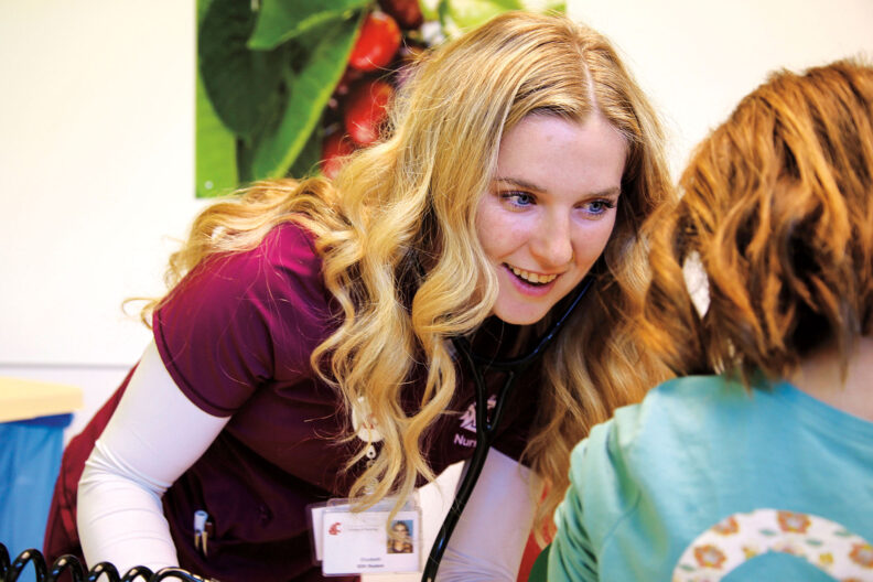 Healthcare student wearing burgundy scrubs uses a stethoscope while interacting with a child in a clinical setting.