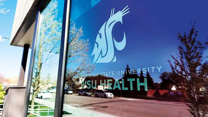 Exterior window displaying the Washington State University Health logo and name, with reflections of trees, parked cars, and nearby buildings