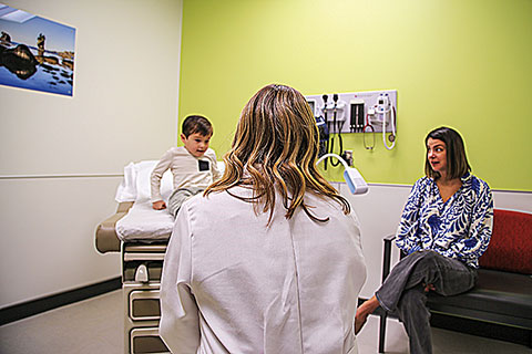 Healthcare provider in a white coat meets with a child seated on an exam table while another adult sits nearby in a medical examination room.