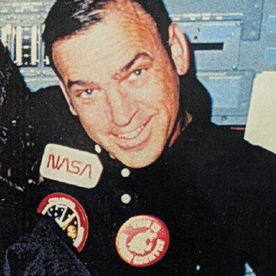 Historic photograph of a man wearing a NASA mission jacket featuring a Washington State University patch, seated inside a spacecraft.