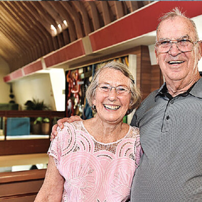 An older woman and an older man stand together inside a building with curved wooden ceiling beams and seats visible in the background.