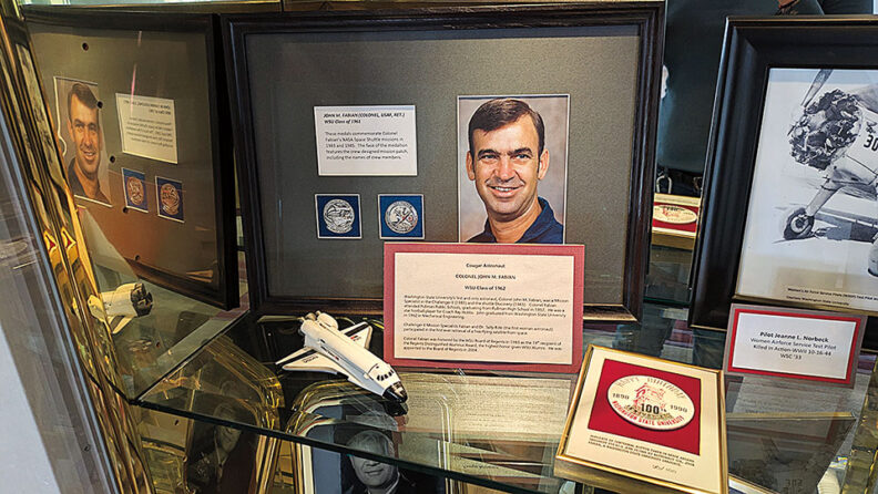 Display case featuring memorabilia related to a Washington State University alumnus, including framed photographs, commemorative plaques, mission patches, and a model space shuttle.
