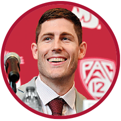 Head-and-shoulders photo of a man speaking at a press conference, seated beside a microphone with a red backdrop displaying the Pac-12 logo.
