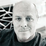 Black-and-white headshot of a man looking toward the camera, with structural beams visible in the background.