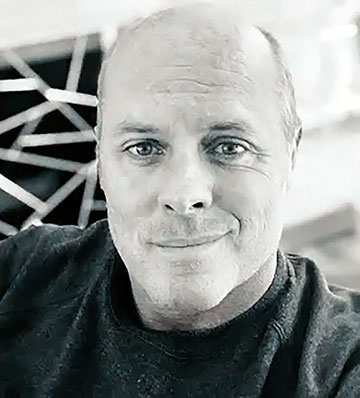 Black-and-white headshot of a man looking toward the camera, with structural beams visible in the background.
