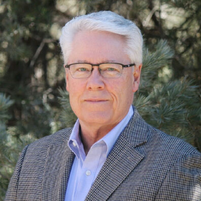 Outdoor portrait of a man with white hair and glasses wearing a patterned blazer and light-colored shirt, standing in front of evergreen trees