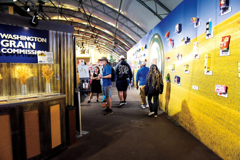 Visitors walk through an agricultural exhibit featuring the Washington Grain Commission, displays of wheat varieties, and products made from Washington-grown crops.