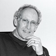 Black-and-white portrait of a man wearing glasses, resting a hand against the chin while seated against a plain background.