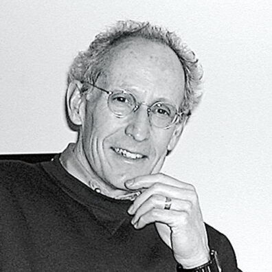 Black-and-white portrait of a man wearing glasses, resting a hand against the chin while seated against a plain background.