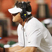 Man wearing a white shirt, visor, and headset seated on the sidelines during a sporting event