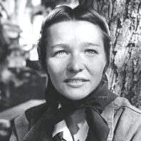 Black-and-white portrait of a young woman seated beside a tree, wearing a neck scarf and jacket, with an outdoor setting and blurred figures in the background.