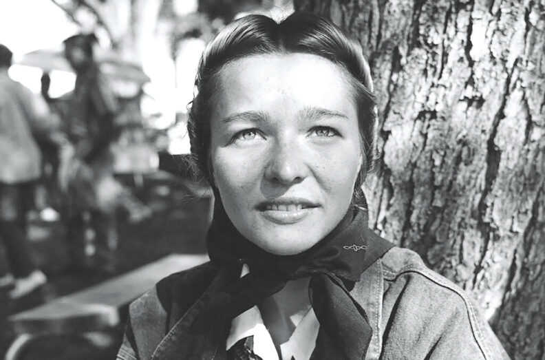 Black-and-white portrait of a young woman seated beside a tree, wearing a neck scarf and jacket, with an outdoor setting and blurred figures in the background.
