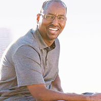 Portrait of a man wearing glasses and a gray polo shirt, seated outdoors against a bright, softly blurred background.
