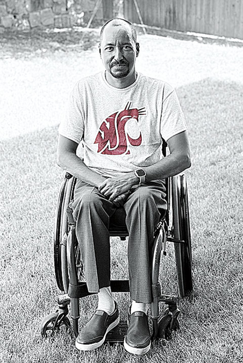 Black-and-white portrait of a man seated in a wheelchair on a lawn, wearing a white T-shirt featuring a crimson Washington State University Cougar logo.