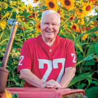 Man wearing a crimson Washington State University football jersey with the number 22 stands behind a red tractor surrounded by tall blooming sunflowers.