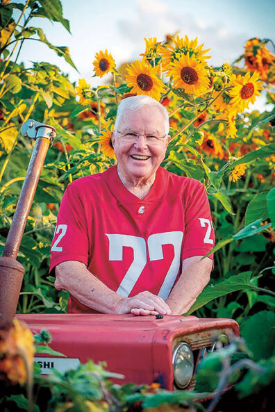 Man wearing a crimson Washington State University football jersey with the number 22 stands behind a red tractor surrounded by tall blooming sunflowers.