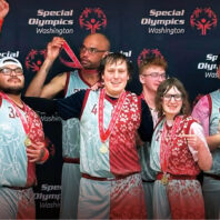 Special Olympics Washington athletes wearing team uniforms pose with medals in front of a Special Olympics Washington backdrop, celebrating after a competition.