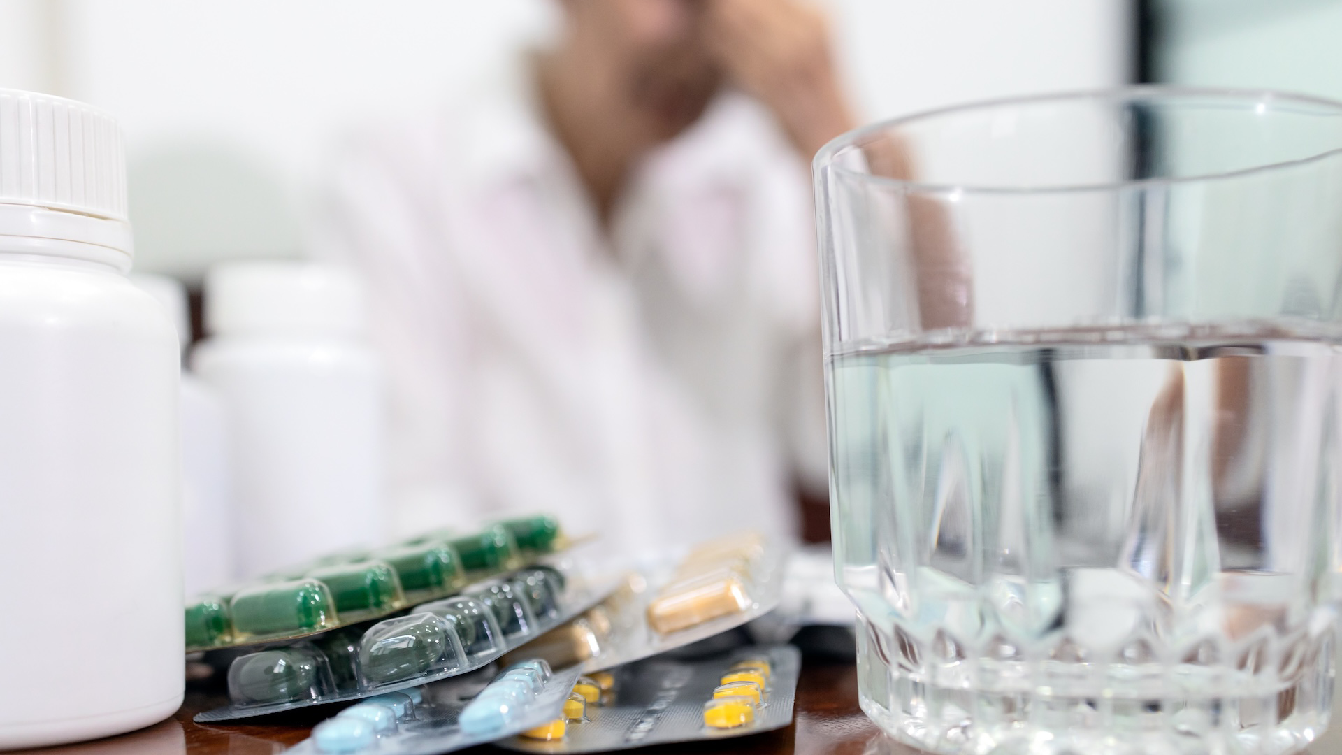 Packages of pills with a glass of water with a distressed person in the background.