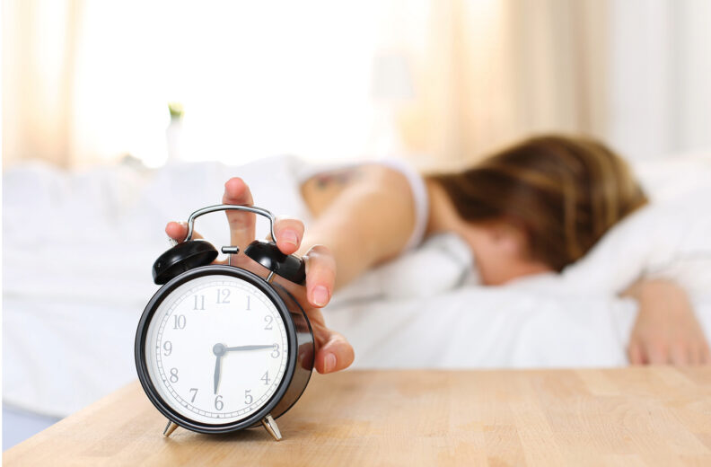 Person laying in bed and reaching out to an alarm clock.