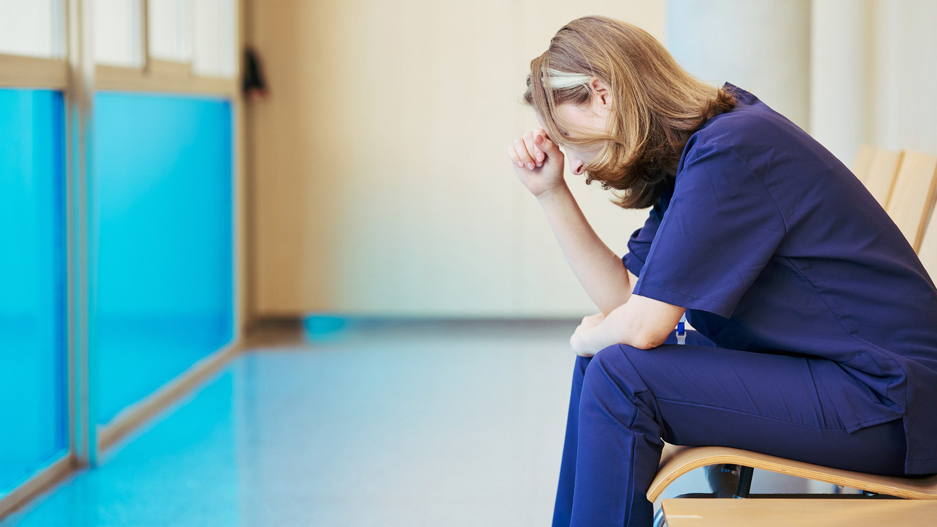 Person sitting in a hallway fatigued, leaning over with thier head on their hand.