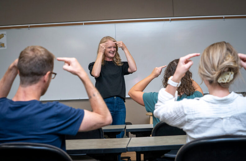 In a classroom with a teacher in front of three students, teaching them ASL signs.