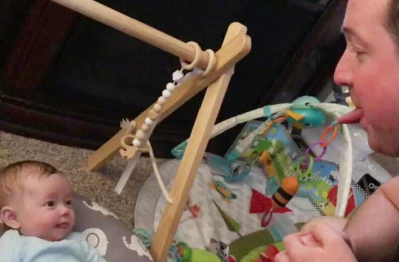 A baby laying on their back making eye contact with their dad who is sticking their tongue out.