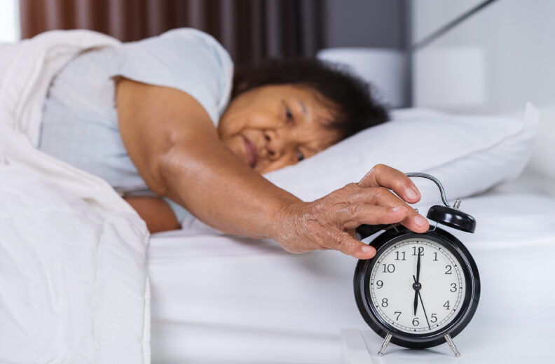 An older person laying in bed, reaching over to an alarm clock.
