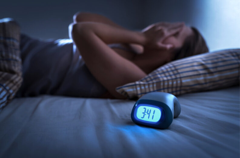 Women laying in bed with her hands over her face as a clock is on the bed showing the time as 3:41 a.m.