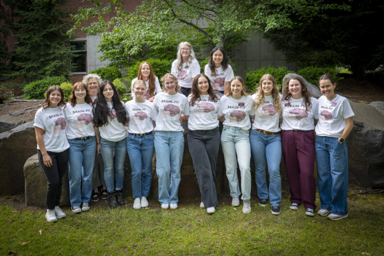 Group of fourteen people outdoors wearing matching white 'BRAIN-FIT' t-shirts with a brain graphic, standing and kneeling in front of large rocks and trees, with greenery and a building in the background.