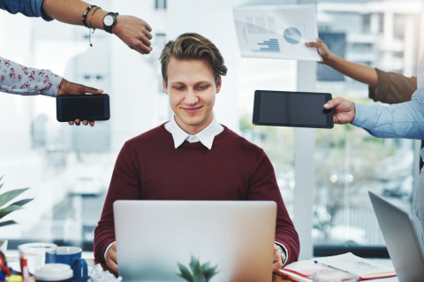 A person working on a laptop at a desk, surrounded by three other individuals holding various devices—a smartphone, a tablet, and a paper with bar graphs.