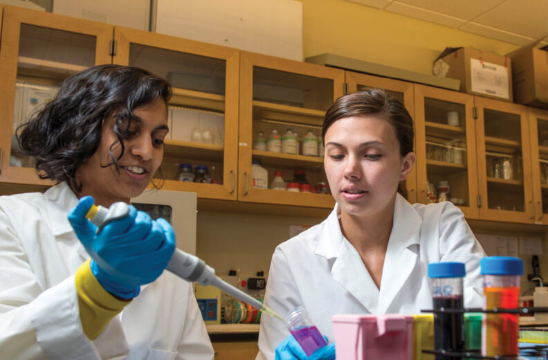 Two people wearing white lab coats work in a laboratory. One person uses a pipette to transfer liquid into a small container, while the other observes. On the counter are colorful vials in a rack and other lab equipment. Shelves in the background hold bottles and supplies.