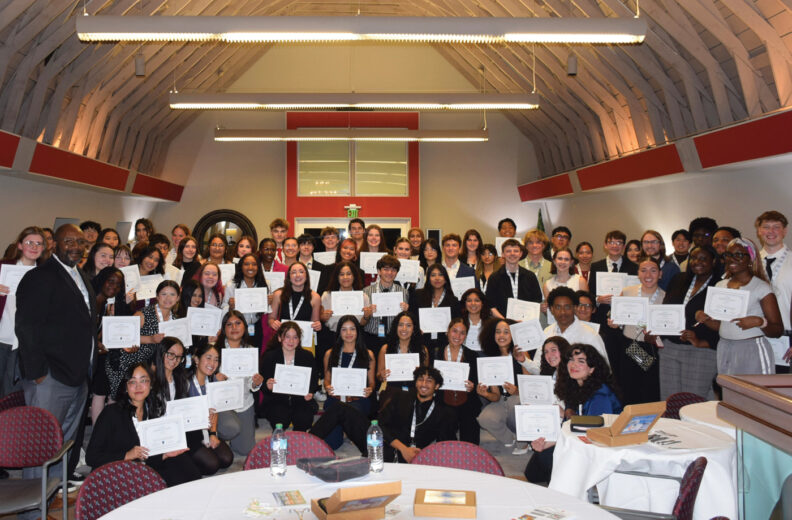 A large group of people pose for a photo in a spacious room with arched ceilings and modern lighting, each holding a certificate. They are arranged in rows, standing and seated, suggesting a formal event or recognition ceremony. Round tables with white tablecloths and items like water bottles and boxes are visible in the foreground.