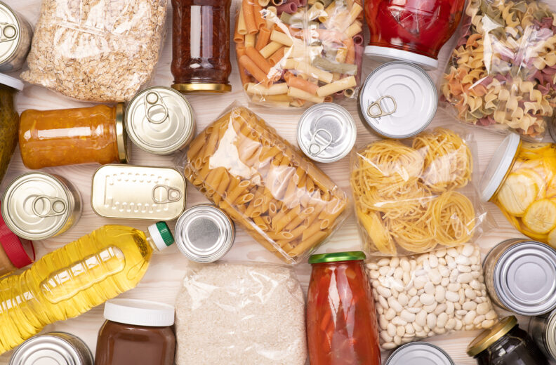 Assorted non-perishable food items arranged on a light wooden surface, including canned goods, jars of sauces, bottles of oil, bags of pasta, rice, beans, and oats.