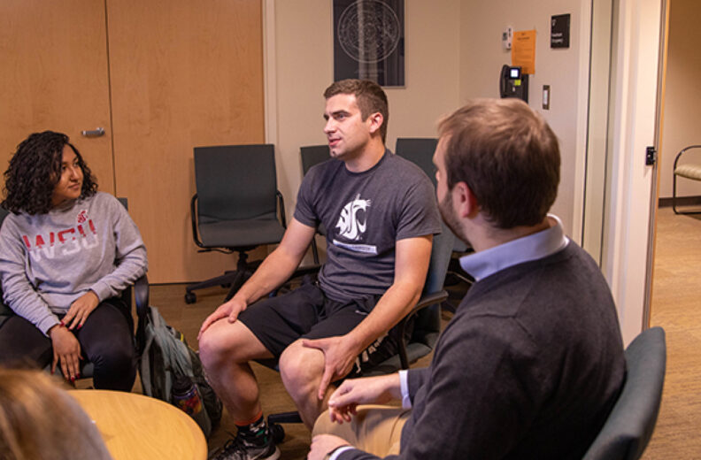 Three people seated in a small room having a conversation. One person wears a gray 'WSU' sweatshirt, another a dark gray t-shirt with a logo, and a third a dark sweater over a light blue shirt. The fourth person is partially visible from behind. The room has beige walls, wooden doors, and office equipment on the wall.