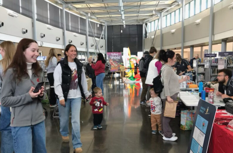 People walk through a bright, open indoor space with high ceilings during what appears to be a community event or health fair. Several tables are set up along the right side with informational materials and staff assisting attendees. Children are present, including one holding a paper bag and another standing near an adult. Colorful signs and displays are visible in the background.