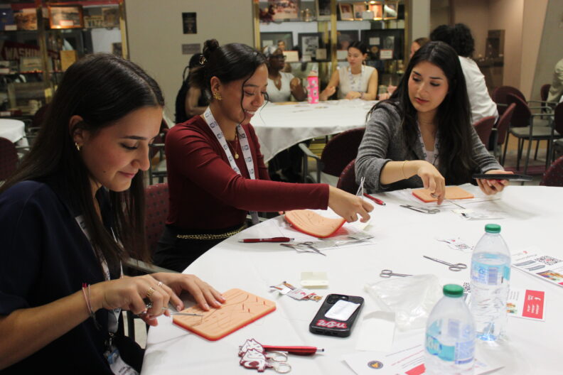 Three people sit around a table practicing suturing techniques on medical training pads, surrounded by tools like scissors, thread, and water bottles.