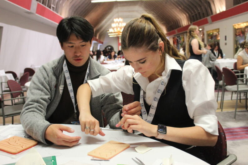 Two individuals seated at a table engage in a hands-on medical training activity. One observes while the other practices suturing on a training pad.