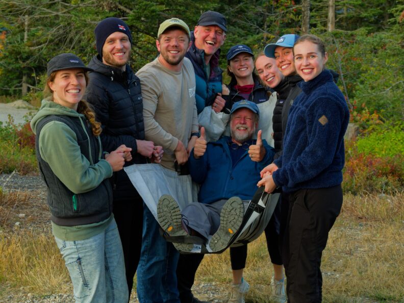Eight people standing on a gravel path outdoors with trees in the background, smiling while holding one person horizontally using a piece of fabric.