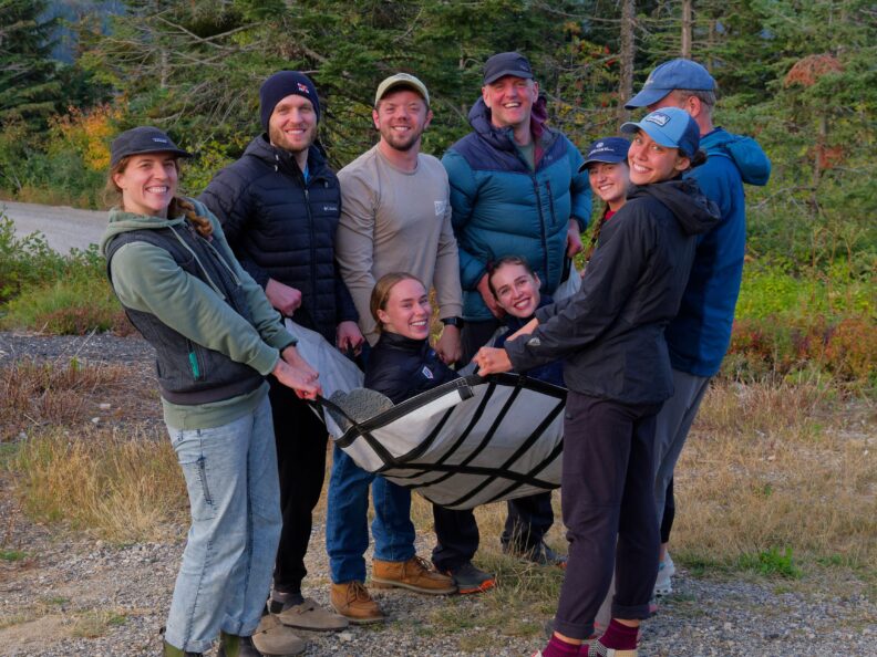 Eight people standing on a gravel path outdoors with trees in the background, smiling while holding two people horizontally using a piece of fabric.