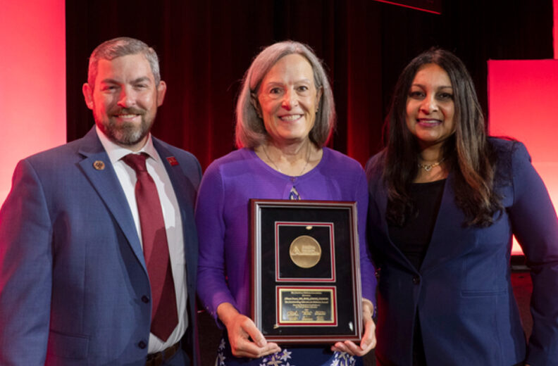 Three individuals standing on a stage with a red and black backdrop. The person in the center is holding a framed award plaque featuring a circular gold medallion and engraved text. The individuals are dressed in formal attire, including suits and a purple top.