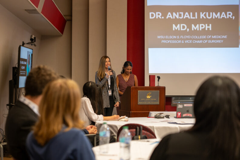 Two speakers stand at a podium presenting to an audience. A screen behind them displays information about Dr. Anjali Kumar, MD, MPH, and her affiliation with WSU Elson S. Floyd College of Medicine.