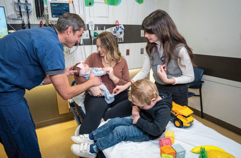 A healthcare professional in scrubs interacts with a baby being held by an adult seated on a hospital bed. Another person with a stethoscope stands nearby, observing. A young child sits on the bed next to colorful toys, including a yellow toy dump truck and stacking blocks. Medical equipment and monitors are visible on the wall in the background.