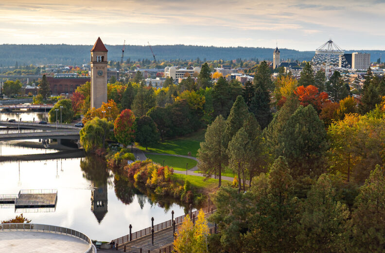 Scenic view of downtown Spokane in autumn, featuring a tree-lined riverfront park with vibrant fall foliage. A red-roofed clock tower stands near the river, with bridges and walking paths crossing the water. City buildings and a large tent-like structure are visible in the background under a partly cloudy sky.